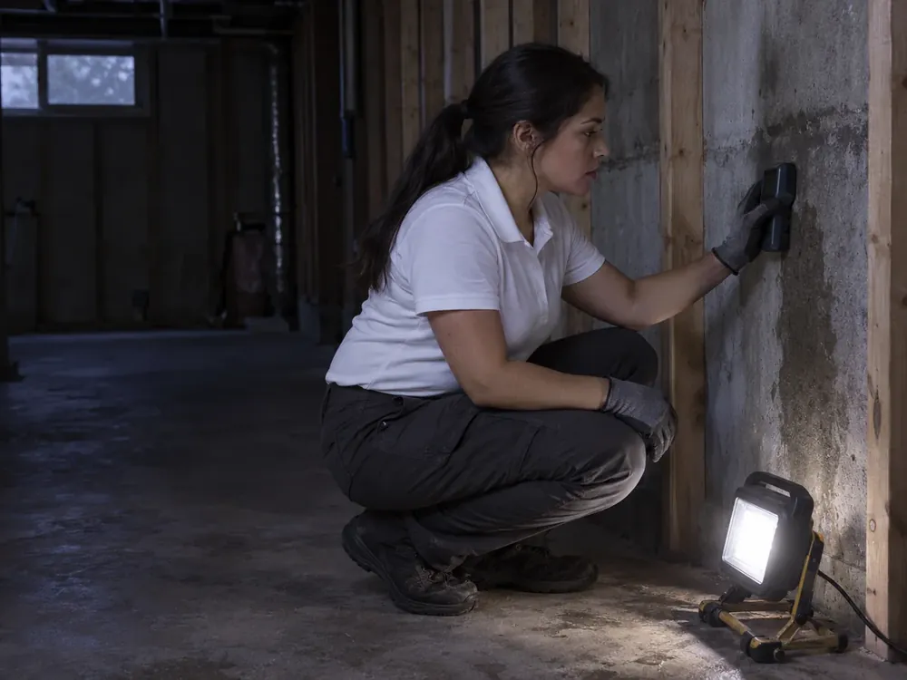 A technician in gloves holds a moisture meter against a damp basement wall by a work lamp